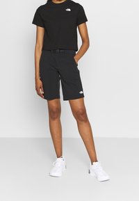 Černé zkrácené tričko a černé šortky, obojí s logem The North Face. Model nosí bílé tenisky. Jednoduché, funkční venkovní oblečení.