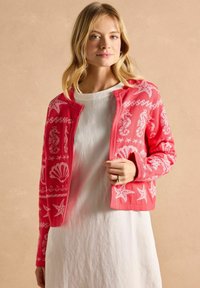 Korálový kardigan s bílými vzory mušlí a hvězdic, dlouhými rukávy a otevřeným předním designem, vrstvený přes lehké bílé šaty.