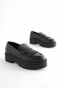 Black leather loafers with a textured finish, featuring a thick, rubber platform sole and a decorative fringe detail on the upper.