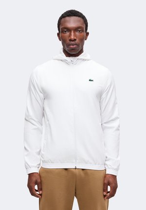 Homme portant une veste blanche zippée avec un petit logo crocodile vert sur la poitrine et un pantalon beige, debout devant un fond uni.
