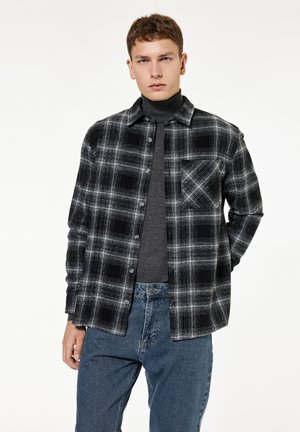 Junger Mann trägt ein schwarz-weiß kariertes Hemd über einem grauen Rollkragenpullover, kombiniert mit blauen Jeans, stehend vor weißem Hintergrund.