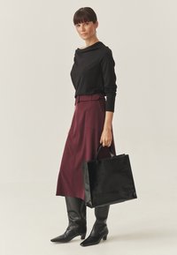 Zwarte top met lange mouwen en een gedrapeerde halslijn, bordeauxrode rok met zakken en zwarte leren laarzen tot aan de knie. Gladde zwarte handtas.