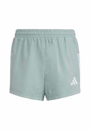 Joven de pie con las manos detrás de la espalda, vistiendo camiseta blanca de Adidas, pantalones cortos gris claro, calcetines blancos y zapatillas blancas sobre fondo blanco.