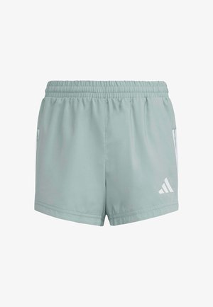 Joven de pie con las manos detrás de la espalda, vistiendo camiseta blanca de Adidas, pantalones cortos gris claro, calcetines blancos y zapatillas blancas sobre fondo blanco.