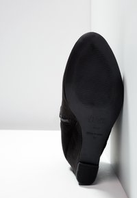 Black suede ankle boot with a low heel, side zipper closure, textured sole, and smooth finish. Branding visible on the sole's underside.