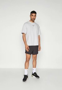 Vyras stovi apsivilkęs šviesiai pilką didelį marškinėlį, juodus šortus, baltas Puma kojines ir juodus sportinius batus paprastoje aplinkoje.