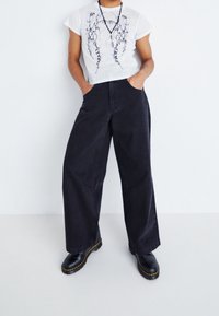 Černé volné džíny s vysokým pasem, spárované s bílým tričkem s grafikou a černým potiskem. Modelka má na sobě černé boty s žlutým švem.