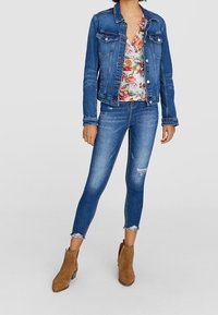 Chaqueta de mezclilla, blusa floral, jeans skinny azules desgastados y botines marrones con detalles de tachuelas. Presenta un ajuste clásico y detalles desgastados.
