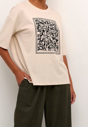 Osoba nosząca beżowy oversize T-shirt z czarno-białym abstrakcyjnym kwadratowym wzorem i luźne ciemnozielone spodnie, jedna ręka w kieszeni.