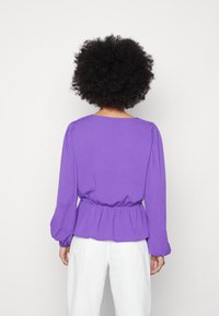 Blusa roxa de manga longa com um padrão de riscas texturizadas, cintura franzida e mangas bufantes; combinada com calças brancas.