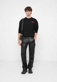 Melna garām piedurknēm krekls ar sarkanu "DIESEL" logotipu, kombinēts ar melnām ādas kravas biksēm ar vairākām kabatām un melnām sporta kurpēm.