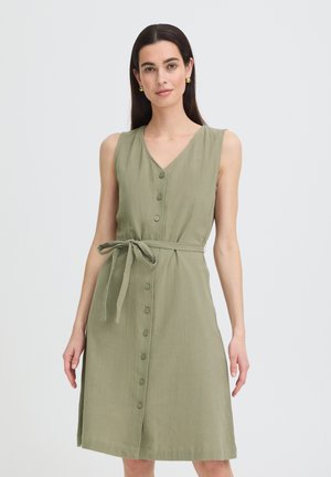 Femme portant une robe sans manches longueur genou vert olive avec boutons sur le devant et ceinture nouée à la taille, debout devant un fond clair uni.