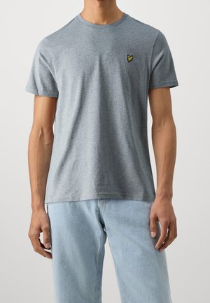 Mann trägt ein schlichtes graues T-Shirt mit kurzen Ärmeln und einem kleinen gelben Vogel-Logo auf der linken Brust, kombiniert mit hellblauen Jeans.