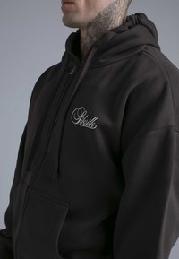 Svart huvtröja i mjukt tyg med framficka, dragsko och en silverfärgad logga på vänster bröst. Mjuk textur och avslappnad passform.