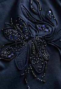 Tissu marine avec un design de fleurs brodées complexes, orné de perles et de paillettes de tailles variées, créant des détails texturés et un éclat.