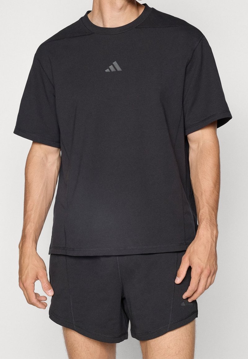 Homme portant un T-shirt de sport noir et un short avec de petits logos Adidas gris sur la poitrine et la jambe gauche du short.