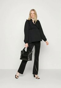 Mulher sorridente, vestindo uma blusa preta com babados, calças de couro pretas, sandálias de salto pretas, segurando uma bolsa preta texturizada, em pé contra um fundo simples.