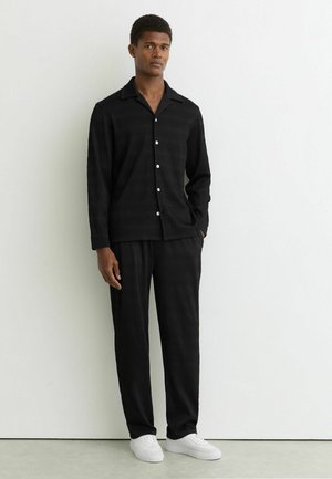 Ung mand står afslappet mod en hvid væg iført en sort tekstureret skjorte med knapper, matchende bukser og hvide sneakers.