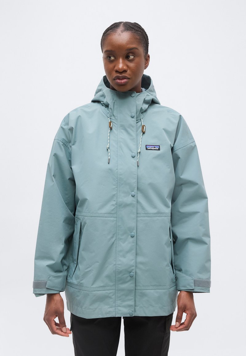 Γυναίκα που φοράει ανοιχτό μπλε αδιάβροχο Patagonia με κουκούλα, στέκεται μπροστά σε απλό λευκό φόντο, κοιτώντας προς τα αριστερά της.