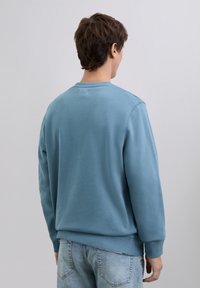 Hombre vestido con un sudadera azul claro y jeans azules desgastados, de espaldas a la cámara, contra un fondo liso.