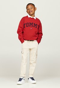 Felpa rossa con logo "TOMMY", camicia bianca con colletto sotto, pantaloni cargo beige e sneakers bianche con accenti blu navy.