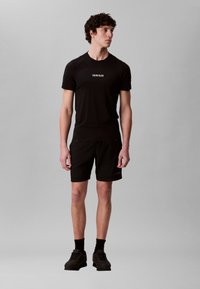 Ung mann vendt mot venstre, iført svart Calvin Klein t-skjorte, svarte shorts, svarte sokker og svarte joggesko mot en ensfarget grå bakgrunn.