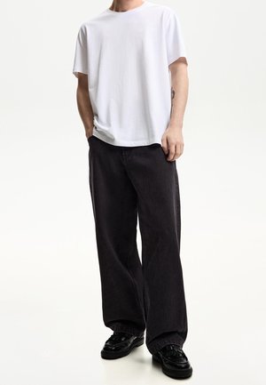 Persoon draagt een losse witte korte mouw t-shirt, brede zwarte broek en glanzende zwarte loafers, staand met één hand in de zak.
