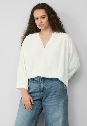 Blouse blanche ample avec un décolleté en V et des manches longues, associée à un jean bleu taille haute. Texture du tissu lisse.