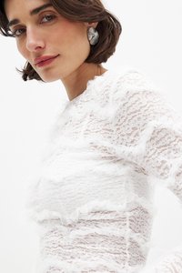 Top bianco in pizzo con un design testo e stratificato, arricchito da dettagli fru-fru. Orecchini circolari argentati completano l'outfit. Tono di pelle uniforme e liscio.