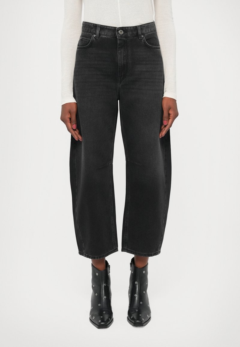 Femme portant un jean noir large taille haute à jambes larges et raccourci avec fermeture par boutons, accompagné de bottines noires à œillets métalliques.
