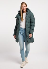 Abrigo acolchado largo y verde oscuro con capucha, presenta un cierre frontal de cremallera y botón a presión. Llevado sobre una camisa clara y jeans azules.