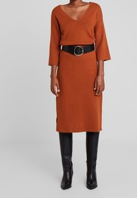 Vestito a maglia color ruggine con scollatura a V, maniche a tre quarti, spacco laterale, abbinato a una cintura nera con motivo coccodrillo e stivali neri al ginocchio.