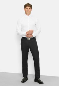 Weißes Langarmhemd mit traditionellem Kragen, kombiniert mit schwarzen Slim-Fit-Hosen und schwarzen Schuhen, stehend vor einem schlichten Hintergrund.