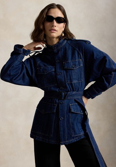 Frau trägt eine dunkelblaue Jeansjacke mit Gürtel und vier vorderen Taschen sowie schwarze Sonnenbrille, posiert mit einer Hand am Nacken und der anderen an der Hüfte.