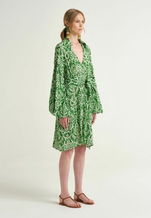 Femme debout portant une robe verte à motifs longueur genou avec manches longues, une ceinture à la taille, des boucles d'oreilles à pompons colorés et des sandales marron.