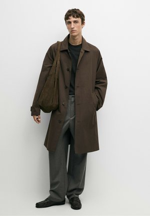 Giovane uomo che indossa un cappotto marrone scuro, pantaloni grigi e scarpe nere con una grande borsa a tracolla marrone, in piedi davanti a uno sfondo bianco uniforme.