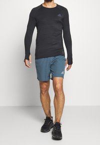 Czarna, długosleeve'owa, sportowa bluzka z teksturowanej tkaniny i otworami na kciuki, w połączeniu z niebieskimi szortami z logo Adidas oraz czarnymi butami sportowymi.