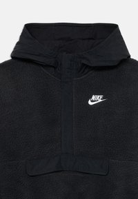 Melnā Nike vilnas pulksteņmala ar pus-krūšu rāvējslēdzēju, augstu apkakli un baltu izšūtu Nike logo uz krūšu.