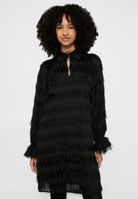 Μαύρο φόρεμα με υφή ζιγκ ζαγκ, μακριά μανίκια με βολάν στις μανσέτες και ντεκολτέ με κλειδί. Κατασκευασμένο από διάφανο ύφασμα με διακοσμητικές φούντες.