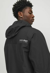 Black hooded jacket made of lightweight fabric, featuring a white logo on the back. Simple design with visible seams and a relaxed fit.