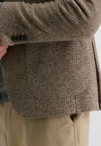 Braun-karierter Blazer mit vier schwarzen Knöpfen an den Ärmeln, getragen über beigen Hosen, die die vordere Tasche und die Seitennaht zeigen.