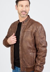 Bruine leren jasje met een gladde afwerking, ritssluiting, geribde manchetten en schouderaccenten. Draagt een zwart shirt eronder.