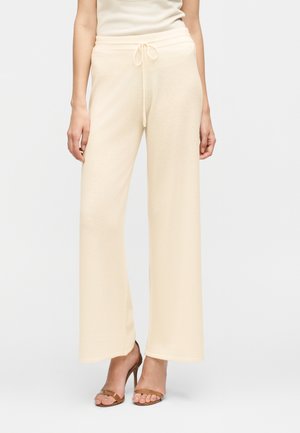 Žena nosi krémové široké pletené nohavice so šnúrkou v páse a hnedé sandále na vysokom podpätku, stojaca pred jednoduchým pozadím.