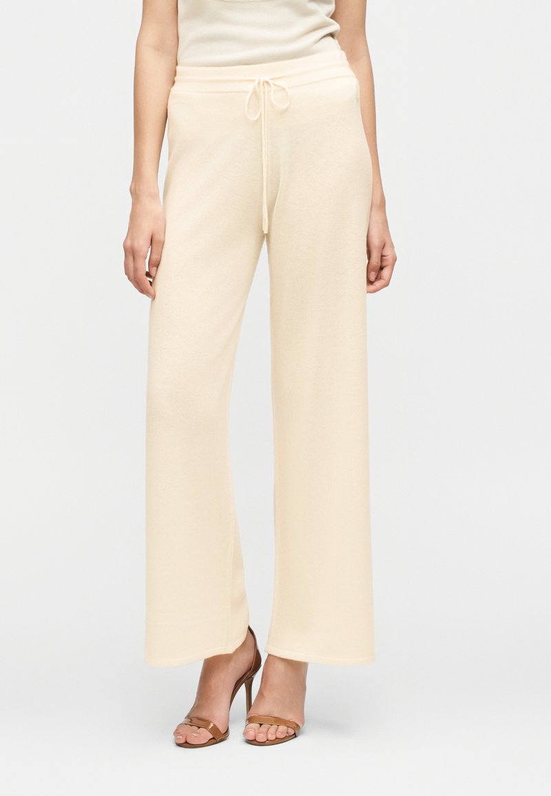 Γυναίκα με κρεμ πλεκτό παντελόνι φαρδιάς γραμμής με κορδόνι στη μέση και καφέ πέδιλα με ψηλό τακούνι, που στέκεται μπροστά σε απλό φόντο.