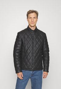 Jachetă puffer neagră, matlasată, cu guler înalt, front cu fermoar și panouri texturate. Purtyată peste un top negru, asortată cu blugi bleumarin.