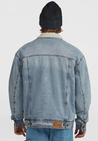Personne portant une veste en denim bleu avec un col en shearling et un bonnet côtelé foncé, vue de dos contre un fond uni.