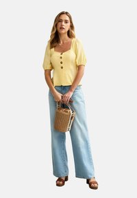 Gul strukturerad blus med korta puff ärmar och bruna knappar, kombinerad med ljusblå vida jeans och en vävd korgväska.