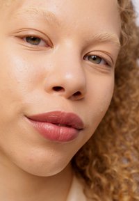 Primer plano del rostro de una persona con cabello rizado, castaño claro, piel clara y labios suaves de un rosa claro. Los ojos son verdes y tienen una expresión natural.