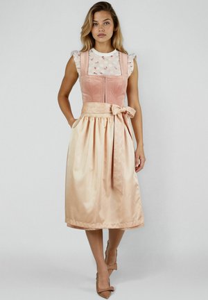 Vrouw draagt een mouwloze roze fluwelen jurk met een bloemmotief blouse eronder en een glanzende beige rok met een strik in de taille.