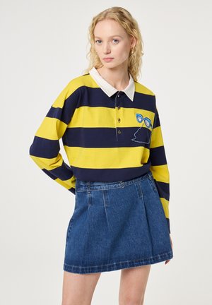 Vrouw met krullend blond haar die een geel en marineblauw gestreept poloshirt met lange mouwen en een blauwe spijkerrok draagt, staand tegen een effen achtergrond.